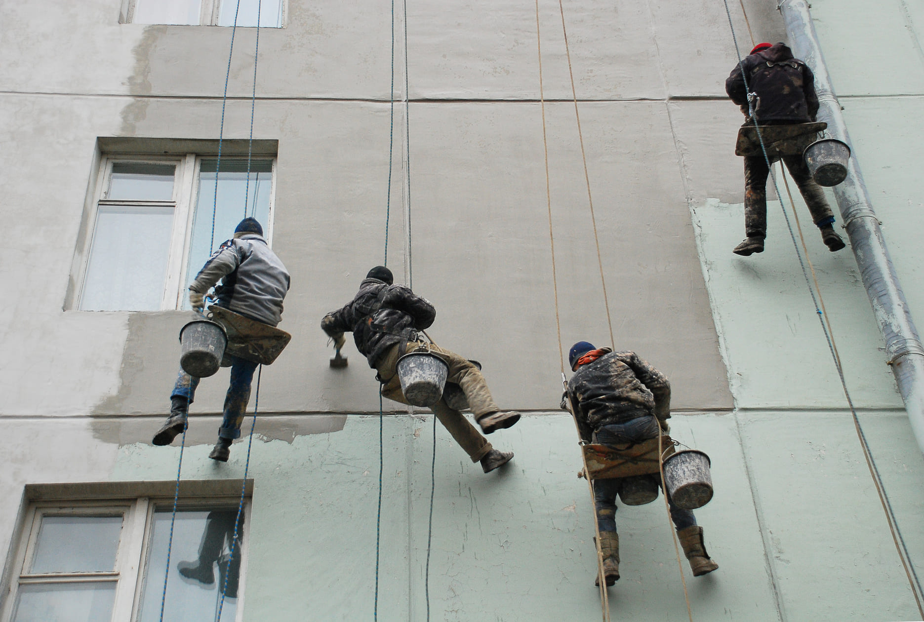 Equipo de trabajadores pintando la fachada de un edificio en altura con cuerdas.